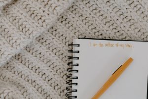 A notebook with an orange pen on top. In orange writing it says "I am the author of my story". The notebook is set against a background of a white knitted woollen blanket