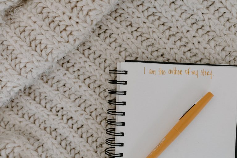 A notebook with an orange pen on top. In orange writing it says "I am the author of my story". The notebook is set against a background of a white knitted woollen blanket